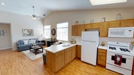 This single-floor home offers a welcoming open-concept layout that seamlessly connects the kitchen and living area, creating a spacious and cohesive living environment. The kitchen is well-appointed with warm honey-toned wood cabinetry, a white appliance suite including a full-size refrigerator, and a peninsula with a clean white countertop that provides additional prep space and serves as a natural divider between the kitchen and living room. Rich hardwood-style flooring runs throughout, adding warmth and continuity to the space. The living area is generously sized and benefits from abundant natural light streaming through multiple windows dressed with classic white blinds. Vaulted ceilings with a ceiling fan enhance the sense of openness and promote airflow, while a skylight above the kitchen adds even more brightness. The living room is comfortably arranged with plush seating, a coffee table, and tasteful décor, lending a cozy yet airy ambiance. The overall design balances practicality with comfort, making this a functional and inviting home suited for everyday living.