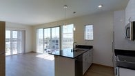 This contemporary single-floor space showcases a clean, modern design with an open-concept layout that seamlessly connects the kitchen and living areas. The kitchen serves as a central feature, anchored by a peninsula with striking dark granite countertops and an undermount sink. Crisp white cabinetry and a stainless steel dishwasher complete the functional workspace, while two stylish cylindrical pendant lights hang overhead, adding a refined touch to the area.

Natural light pours generously into the space through expansive floor-to-ceiling sliding glass doors, offering views of the surrounding cityscape and filling the interior with a bright, airy atmosphere. A secondary window with horizontal blinds provides additional light while allowing privacy control. The light-toned hardwood flooring runs throughout, complementing the neutral white walls and contributing to the space's clean, minimalist aesthetic.

The open living area benefits directly from the natural light and views afforded by the large glass doors, creating a welcoming and versatile environment. Recessed lighting supplements the pendant fixtures, ensuring the space is well-lit at all times. Overall, the unit presents a polished, contemporary feel with thoughtful design details throughout.