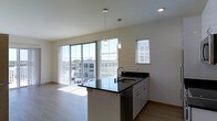 This contemporary single-floor space showcases a clean, modern design with an open-concept layout that seamlessly connects the kitchen and living areas. The kitchen serves as a central feature, anchored by a peninsula with striking dark granite countertops and an undermount sink. Crisp white cabinetry and a stainless steel dishwasher complete the functional workspace, while two stylish cylindrical pendant lights hang overhead, adding a refined touch to the area.

Natural light pours generously into the space through expansive floor-to-ceiling sliding glass doors, offering views of the surrounding cityscape and filling the interior with a bright, airy atmosphere. A secondary window with horizontal blinds provides additional light while allowing privacy control. The light-toned hardwood flooring runs throughout, complementing the neutral white walls and contributing to the space's clean, minimalist aesthetic.

The open living area benefits directly from the natural light and views afforded by the large glass doors, creating a welcoming and versatile environment. Recessed lighting supplements the pendant fixtures, ensuring the space is well-lit at all times. Overall, the unit presents a polished, contemporary feel with thoughtful design details throughout.