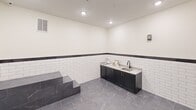 This purpose-built pet grooming room is thoughtfully designed to combine functionality with a clean, polished aesthetic. The space is outfitted with two raised, marble-finish wash stations equipped with coiled hose attachments, ideal for bathing pets of various sizes. A wall-mounted professional pet dryer is conveniently positioned between the stations for efficient use. A dark cabinetry vanity with granite countertops and a utility sink offers dedicated space for grooming supplies and cleanup, complete with gold-toned hardware that adds a refined touch. White subway tile lines the lower half of all walls, capped with a dark contrasting border that transitions to smooth, cream-painted upper walls. The flooring throughout is a large-format grey marble-look tile, lending the room a sophisticated yet durable surface suited to wet environments. Recessed ceiling lights provide bright, even illumination across the entire room. Charming paw print wall decals and framed pet-themed artwork contribute a welcoming, playful atmosphere. A paneled door with an automatic closer ensures secure and easy access. This well-appointed grooming room is a standout amenity, balancing practical utility with stylish design.