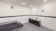 This purpose-built pet grooming room is thoughtfully designed to combine functionality with a clean, polished aesthetic. The space is outfitted with two raised, marble-finish wash stations equipped with coiled hose attachments, ideal for bathing pets of various sizes. A wall-mounted professional pet dryer is conveniently positioned between the stations for efficient use. A dark cabinetry vanity with granite countertops and a utility sink offers dedicated space for grooming supplies and cleanup, complete with gold-toned hardware that adds a refined touch. White subway tile lines the lower half of all walls, capped with a dark contrasting border that transitions to smooth, cream-painted upper walls. The flooring throughout is a large-format grey marble-look tile, lending the room a sophisticated yet durable surface suited to wet environments. Recessed ceiling lights provide bright, even illumination across the entire room. Charming paw print wall decals and framed pet-themed artwork contribute a welcoming, playful atmosphere. A paneled door with an automatic closer ensures secure and easy access. This well-appointed grooming room is a standout amenity, balancing practical utility with stylish design.