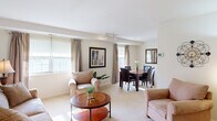 This single-floor residence offers a practical and comfortable layout comprising one bedroom, one bathroom, a combined living and dining room, a kitchen, and two closets. The open-plan living and dining area is bright and welcoming, featuring large windows dressed with striped drapes and roller shades that allow generous natural light to fill the space. Neutral-toned carpet, clean white walls, and track lighting lend the area a warm, airy ambiance. The dining space accommodates a full-sized table and chairs, accented by a decorative mirror and framed artwork. A cozy breakfast nook adjacent to the kitchen features a glass-top bistro table with wrought-iron chairs, separated from the main living area by a charming interior window partition. The kitchen is equipped with warm oak cabinetry, white countertops, a tile backsplash, and a gas range. The spacious bedroom features plush carpeting, a large window for natural light, and direct sightlines into the living area. Two closets provide ample storage. The full bathroom includes a tiled tub-shower combination. Throughout, the space maintains a cohesive, neutral palette that creates a calm and functional living environment.