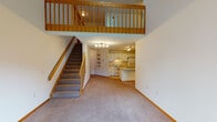 This two-story space is defined by its open, airy design and thoughtful use of vertical space. On the main floor, a generously sized living area features soaring ceilings that extend upward to the second level, where a wood-railed loft overlooks the space below. Soft, neutral carpeting runs throughout the living areas, complemented by warm wood trim and baseboards. The kitchen is positioned adjacent to the living space and features white cabinetry, light countertops, a full suite of white appliances including a range and refrigerator, and a peninsula that provides additional workspace. Built-in open shelving on a nearby wall adds functional display space. A large window on the main floor allows natural light to pour in, offering views of the surrounding outdoor greenery. The upper level opens as a versatile loft space, illuminated by multiple skylights and a ceiling fan, creating a bright and comfortable area. Additional rooms on this level include carpeted bedrooms with vaulted ceilings and ceiling fans, one featuring a skylight and closet access. Two bathrooms are present, one with a wide vanity and tub, and another with a walk-in glass-door shower, both finished with light tile flooring and ample cabinetry.