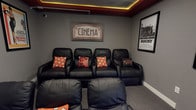 This single-floor home cinema room is thoughtfully designed to deliver an authentic movie theater experience. The space is anchored by multiple rows of plush black leather recliner seats, each equipped with armrests and accented with themed decorative pillows featuring popcorn and film-related motifs. A tiered seating arrangement ensures clear sightlines to the large wall-mounted flat-screen television at the front of the room, which serves as the primary viewing display.

The ceiling is a standout architectural feature, finished in deep red with integrated recessed lighting and warm amber LED strip lighting that runs along the perimeter, casting a dramatic, cinema-like glow throughout the space. The walls are painted in a muted grey tone, reinforcing the immersive, darkened atmosphere typical of a professional screening room.

Movie memorabilia adds considerable character to the space, including a prominent "Cinema" marquee-style sign and a framed Rocky movie poster. Floating wall shelves below the screen house an extensive collection of DVDs, further emphasizing the room's dedication to film. The overall ambiance is intimate and purpose-built, combining comfortable seating, controlled lighting, and curated décor to create a private cinema environment ideal for dedicated movie viewing.