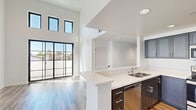 This contemporary two-story residence showcases a clean, modern aesthetic throughout, highlighted by an open and airy layout filled with natural light. The main living area impresses with dramatically high ceilings and large black-framed floor-to-ceiling sliding doors and clerestory windows that flood the space with sunlight and provide views of the surrounding neighborhood. Wood-look flooring flows seamlessly throughout both levels, complementing the crisp white walls and minimalist design.

The kitchen is thoughtfully appointed with dark charcoal shaker-style cabinetry, white quartz countertops, stainless steel appliances including a refrigerator, range, and built-in microwave, and a peninsula with a stainless steel sink — ideal for both everyday cooking and entertaining. The open layout connects the kitchen effortlessly to the main living area.

Bedrooms are generously proportioned, with wide windows framing neighborhood views and recessed lighting overhead. The primary suite includes a spacious walk-in closet outfitted with custom white shelving, hanging rods, and built-in drawers. The primary bathroom features a dual-sink vanity with white cabinetry, a large mirror, and wood-look flooring that adds warmth to the clean, white space.

Outdoors, the property features private exterior areas enclosed by white stucco walls, set within a Mediterranean-style neighborhood with terracotta rooftops and mature palms nearby.