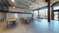 Located at 695 S Santa Fe Ave in Los Angeles' vibrant Arts District, this single-floor commercial space showcases a compelling industrial aesthetic throughout. The open-plan layout is anchored by polished concrete floors, exposed concrete ceilings with visible ductwork and piping, and striking raw brick accent walls that lend an authentic warehouse character to the environment.

The main gathering area is generously proportioned, accommodating multiple rows of sturdy wooden communal tables paired with industrial-style metal stools. Pendant lighting hangs from the ceiling at varying heights, providing warm, focused illumination across the seating zones. Large floor-to-ceiling windows and oversized glass garage-style doors flood the space with abundant natural light while offering open street views, connecting the interior to the surrounding neighborhood.

A secondary seating area mirrors this design language, featuring cinder block walls and additional large-format windows that maintain the bright, airy atmosphere. A dedicated service or catering area is outfitted with an extended run of light wood cabinetry, open shelving, stainless steel sinks, and ample counter workspace — well-suited for food preparation or event catering needs. Together, these elements create a versatile, functional space ideal for events, gatherings, or collaborative work.