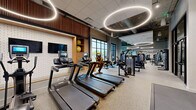 This contemporary fitness center offers a comprehensive, well-organized workout environment across a generously sized single-floor layout. The space features an industrial-modern aesthetic, with exposed ductwork and concrete ceilings complemented by striking circular halo pendant lights that illuminate the facility with a warm, energizing glow. Large black-framed windows along the perimeter flood the space with natural light and provide views to the outdoors.

The cardio zone is equipped with multiple treadmills, stationary bikes, and elliptical machines, positioned alongside a built-in media wall with a flat-screen TV and open shelving for stability balls. Adjacent to this area, a dedicated strength training section houses a full lineup of Precor selectorized machines and cable systems, catering to a wide range of muscle groups.

A free weights area features a complete dumbbell rack alongside adjustable benches and mirrors that enhance the sense of space. A functional training zone includes a bold yellow multi-purpose rig with a heavy bag, resistance attachments, and wall-mounted bars. A stretching and floor exercise area is defined by thick mat flooring and accented by a dramatic abstract mural. Glass partition walls divide select zones, maintaining an open yet organized feel throughout the facility.