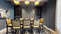 This elegantly appointed space exudes a refined, upscale ambiance through a carefully curated combination of materials and furnishings. The focal point of the room is a striking exposed brick accent wall in a cool grey tone, flanked on either side by dark wood diamond-pattern wine racks that extend from floor to ceiling, offering both functional storage and dramatic visual appeal. A long communal dining or bar-height table anchors the space, surrounded by luxurious quilted gold leather chairs with dark metal frames and decorative ring hardware, lending a glamorous yet structured aesthetic. Overhead, a pair of ornate fringe chandeliers with gold metallic tassels cast a warm, atmospheric glow throughout the room. The deep blue-grey walls complement the rich tones of the furnishings, while a detailed patterned carpet in blue and white tones adds visual depth underfoot. The overall design balances industrial elements, such as the brick wall and dark metal accents, with opulent touches in the gold upholstery and statement lighting, creating a space that feels both inviting and sophisticated — well-suited for intimate dining or social gatherings.