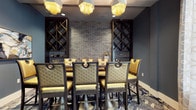 This elegantly appointed space exudes a refined, upscale ambiance through a carefully curated combination of materials and furnishings. The focal point of the room is a striking exposed brick accent wall in a cool grey tone, flanked on either side by dark wood diamond-pattern wine racks that extend from floor to ceiling, offering both functional storage and dramatic visual appeal. A long communal dining or bar-height table anchors the space, surrounded by luxurious quilted gold leather chairs with dark metal frames and decorative ring hardware, lending a glamorous yet structured aesthetic. Overhead, a pair of ornate fringe chandeliers with gold metallic tassels cast a warm, atmospheric glow throughout the room. The deep blue-grey walls complement the rich tones of the furnishings, while a detailed patterned carpet in blue and white tones adds visual depth underfoot. The overall design balances industrial elements, such as the brick wall and dark metal accents, with opulent touches in the gold upholstery and statement lighting, creating a space that feels both inviting and sophisticated — well-suited for intimate dining or social gatherings.