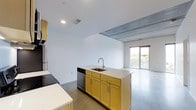 This single-floor space showcases a thoughtfully designed open-concept layout that seamlessly connects the kitchen and living areas. The kitchen is a standout feature, centered around a freestanding island with a stainless steel sink, light countertops, and warm yellow-toned wood cabinetry that adds a welcoming contrast to the otherwise neutral palette. A stainless steel dishwasher is integrated beneath the island, while additional cabinetry and a refrigerator line the perimeter, providing generous storage and functionality.

The flooring throughout is polished concrete, lending an industrial yet refined character to the space. This aesthetic is further reinforced by an exposed concrete beam detail at the ceiling transition point between the kitchen and living area, adding architectural interest and depth. Recessed lighting illuminates the kitchen zone, complementing the natural light that filters in through a sliding glass door in the adjacent living area. The door opens to what appears to be an outdoor space, enhancing the sense of openness. Crisp white walls keep the overall ambiance clean, bright, and versatile.
