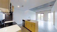 This single-floor space showcases a thoughtfully designed open-concept layout that seamlessly connects the kitchen and living areas. The kitchen is a standout feature, centered around a freestanding island with a stainless steel sink, light countertops, and warm yellow-toned wood cabinetry that adds a welcoming contrast to the otherwise neutral palette. A stainless steel dishwasher is integrated beneath the island, while additional cabinetry and a refrigerator line the perimeter, providing generous storage and functionality.

The flooring throughout is polished concrete, lending an industrial yet refined character to the space. This aesthetic is further reinforced by an exposed concrete beam detail at the ceiling transition point between the kitchen and living area, adding architectural interest and depth. Recessed lighting illuminates the kitchen zone, complementing the natural light that filters in through a sliding glass door in the adjacent living area. The door opens to what appears to be an outdoor space, enhancing the sense of openness. Crisp white walls keep the overall ambiance clean, bright, and versatile.