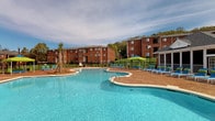 This inviting outdoor amenity area centers around a generously sized, freeform swimming pool with crystal-clear blue water, designed to evoke a resort-style atmosphere. The pool is complemented by an expansive stamped concrete deck that surrounds the water on multiple sides, providing ample space for relaxation and recreation. Numerous blue-and-white lounge chairs are arranged across the deck, offering residents plenty of spots to unwind, sunbathe, or socialize. Shaded areas with vibrant green canopy umbrellas and curtained cabanas provide comfortable retreats from the sun.

Adjacent to the pool area is a screened-in clubhouse structure, featuring large screen-panel walls and white railings that allow for unobstructed views of the pool while offering protection from the elements. Inside, ceiling fans maintain airflow, and the interior is furnished with seating and a kitchen-bar area, making it ideal for gatherings and events. Decorative brick accents tie the clubhouse visually to the surrounding multi-story brick residential buildings, creating a cohesive architectural aesthetic throughout the community.

The entire pool area is enclosed by a black iron fence, ensuring both safety and security. Mature trees and palm-style plantings at the perimeter add a touch of lush greenery, enhancing the resort-like ambiance of this well-appointed community outdoor space.