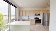 This contemporary single-floor unit offers a clean, modern aesthetic throughout its thoughtfully designed layout. The kitchen is a standout feature, equipped with stainless steel appliances including a refrigerator, electric range, built-in microwave, and dishwasher. Light natural wood cabinetry lines the walls from counter to ceiling, complemented by a white tile backsplash, white countertops, and a central island that provides additional prep and serving space. Large windows flood the kitchen with natural light and offer open views of the surrounding area. The living spaces are finished in crisp white walls with light-toned luxury vinyl tile flooring throughout, creating a bright and airy atmosphere. Multiple rooms feature large windows dressed with dark roller shades, framing sweeping outdoor views of the urban surroundings and green treetops. Baseboard heaters are present throughout, and at least one room is equipped with a wall-mounted mini-split unit for climate control. A dedicated room includes a spacious walk-in closet with built-in shelving and hanging rods. A versatile room with built-in desk cabinetry and a large picture window serves well as a study or workspace. The overall design emphasizes clean lines, natural light, and functional simplicity.