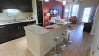 This well-appointed single-floor residence showcases a thoughtfully designed open-concept layout that connects the kitchen and living areas in a seamless, functional flow. The kitchen is outfitted with dark espresso cabinetry, a full-size refrigerator, and a built-in dishwasher, complemented by light-toned hardwood flooring that runs throughout the space. A prominent kitchen peninsula with a granite countertop serves as the centerpiece, offering generous prep space and casual seating with modern acrylic and chrome bar stools — ideal for both everyday dining and entertaining. The living area extends naturally from the kitchen, anchored by a bold accent wall in deep red that adds warmth and visual interest. A white sofa and coordinating furnishings create a clean, contemporary look, while a decorative mirror-panel wall art piece adds a refined touch. Natural light filters in through sizable windows dressed with blinds, and a ceiling fan contributes to the room's comfort. The overall ambiance is modern and inviting, balancing bold design choices with a neutral foundation to create a cohesive, stylish living environment.