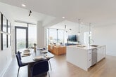 This contemporary single-floor residence showcases a thoughtfully designed open-concept layout that seamlessly connects the kitchen, dining, and living areas. The living room is a standout, anchored by dramatic floor-to-ceiling windows that frame sweeping city and treetop views, flooding the space with natural light. A sculptural multi-arm pendant light adds an artistic focal point overhead.

The chef's kitchen is outfitted with high-gloss white cabinetry reaching to the ceiling, a herringbone-patterned metallic tile backsplash, stainless steel appliances including a gas range and French door refrigerator, and a generous waterfall island with seating. Globe pendant lights add a refined finish.

The dining area features a substantial table with velvet seating beneath a curated gallery wall of black-and-white photography. Light wide-plank hardwood flooring runs throughout, tying each space together cohesively.

The primary bedroom offers its own wall of floor-to-ceiling windows with lush outdoor views, creating a serene retreat. It connects directly to a well-appointed en-suite bathroom featuring a glass-enclosed walk-in shower with white tile surround.

The entry hallway is clean and functional, with louvered closet doors and matte black hardware accents that echo the modern aesthetic carried throughout the entire residence.