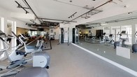 This fitness center is designed to support a comprehensive workout experience within a single, open-plan floor. The space features an industrial-modern aesthetic, with exposed ductwork, pipe systems, and track lighting running along the white-painted ceiling, lending the room a clean yet dynamic character. Durable rubber flooring spans the main exercise area, providing both comfort and safety underfoot.

The cardio zone is equipped with treadmills, elliptical trainers, upright and recumbent bikes, and a rowing machine, many from Precor — all positioned near large windows that bring in ample natural light and views to the exterior. A wall-mounted flat-screen TV serves this section for entertainment during workouts.

The strength and functional training area offers a cable machine, a multi-function cable/pulley station, adjustable benches, a full rack of dumbbells, kettlebells, medicine balls in multiple weights, exercise mats, and stability balls. A dedicated floor space with marked boundaries provides room for stretching and functional movements.

Floor-to-ceiling mirrors line the walls throughout, visually expanding the space and allowing users to monitor their form. A water cooler and waste receptacle are conveniently positioned near the entrance. Signage for fitness center rules is posted on the wall, reflecting an organized and professionally maintained facility.