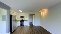 This single-floor space features a clean, modern design with an open-concept layout that seamlessly connects the kitchen and living areas. The kitchen is well-appointed with stainless steel appliances, including a gas range, microwave, and refrigerator, all complemented by crisp white shaker-style cabinetry and stone countertops. A skylight-style recessed fixture brightens the kitchen, while track lighting in the living area provides versatile illumination. Rich, dark hardwood flooring runs throughout the space, offering a striking contrast against the bright white walls and trim. The open living area provides a generous sense of space, enhanced by the clean sightlines flowing from the kitchen toward a hallway that leads to additional rooms beyond. The neutral color palette and contemporary finishes create a bright, airy ambiance that is both inviting and functional. HVAC vents are integrated discreetly into the ceiling, ensuring comfort throughout. The overall design strikes a balance between modern aesthetics and practical living, making this a well-designed and highly functional space.