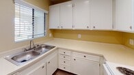 Located at 1401 N Placentia Ave in Fullerton, California, this single-floor unit offers a practical and functional layout across several connected living spaces. The living area is finished with patterned carpeting and warm beige walls, creating a neutral and inviting atmosphere. A wall-mounted air conditioning unit provides climate control, while a sliding glass door leads to an outdoor balcony, allowing natural light to fill the room and offering views of surrounding greenery. A ceiling fan is installed in an adjacent area, enhancing airflow throughout the space.

The kitchen is equipped with white raised-panel cabinetry, offering ample upper and lower storage. Light-toned countertops provide generous prep space, and a double-basin stainless steel sink sits beneath a window fitted with horizontal blinds. Dark hardwood-style flooring in the kitchen and entryway areas contrasts with the carpeted living spaces, adding visual interest and easy-to-clean surfaces in high-traffic zones.

The bathroom features a vanity with a single sink, white cabinetry below, and a mirrored medicine cabinet illuminated by a wall-mounted light fixture. A closet with open shelving is positioned nearby, offering convenient storage. Textured ceilings are consistent throughout the unit, contributing to its classic residential character.