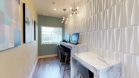 This single-floor space functions as a dedicated business center, thoughtfully designed to balance functionality with modern aesthetics. The room features a long, marble-topped counter workstation that runs along one wall, providing ample workspace for multiple users simultaneously. Several desktop computers are arranged along the counter, accompanied by sleek black bar-height chairs that complement the contemporary style.

One wall is clad in a striking white textured panel with a dimensional, wave-like relief pattern, adding architectural interest and visual depth to the space. The opposing wall is painted in a soft sage green, creating a calming contrast against the crisp white surfaces. A window at the end of the room allows natural light to filter in, offering a view of the surrounding outdoor greenery.

Overhead, a sputnik-style chandelier serves as a focal point, providing warm ambient lighting that enhances the room's modern character. The flooring consists of wood-look planks in a neutral gray tone, contributing to the clean, cohesive design. Together, these elements create a polished and inviting workspace suited for professional tasks.