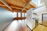 Located at 313 E Willow St in Syracuse, NY, this two-bedroom, one-bathroom loft-style space spans two floors and showcases a striking blend of industrial and modern design. The main level is generously proportioned, featuring soaring wood-planked ceilings supported by exposed steel beams, rich hardwood floors, and dramatic exposed brick walls that run the length of the living area. Multiple large windows flood the space with natural light, enhancing the open, airy feel of the living room. A dedicated entrance leads into the expansive main floor, where the living area flows seamlessly toward the kitchen. The kitchen is efficiently laid out in a galley configuration, equipped with stainless steel appliances including a range and refrigerator, ample white cabinetry, and tile flooring. A skylight above the kitchen adds an additional source of natural light. The primary bedroom continues the industrial aesthetic with exposed brick and wood-beam ceilings. The upper level, accessed via staircase, provides additional bedroom space and closet storage. The bathroom is cleanly finished with a tiled tub-shower combination, a wood-framed vanity mirror, and stone-style tile flooring. Two closets throughout the unit provide practical storage solutions.