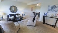 This two-story model home showcases a thoughtfully designed open-concept layout that connects the living and dining areas in a cohesive, functional arrangement. The living area is generously sized, featuring soft wall-to-wall carpeting in a warm neutral tone that extends throughout the space, creating a comfortable and inviting atmosphere. Smooth, light-colored walls provide a clean backdrop, while the layout allows for flexible furniture arrangements to suit a variety of lifestyles.

A defined dining area sits adjacent to the living space, partially set apart by a subtle architectural transition in the ceiling, adding visual interest and a sense of structure without fully closing off the room. A ceiling fixture overhead anchors the dining zone, which comfortably accommodates a dining table and chairs. The open passage between the two areas promotes a natural flow, making the space ideal for both everyday living and entertaining.

The overall aesthetic is bright and neutral, with understated finishes that offer a versatile canvas for personalization. The two-floor layout suggests additional living space above, providing room for bedrooms and additional functional areas beyond the main level.