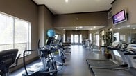 This fitness center offers a comprehensive workout environment designed to accommodate a variety of exercise routines. The space is finished with neutral taupe-toned walls and crown molding details, lending a polished, upscale aesthetic. Recessed ceiling lighting provides consistent illumination throughout, while large windows and glass-paneled doors along the far wall invite natural light deep into the room, creating an open and energizing atmosphere.

The layout is thoughtfully organized, with cardio equipment—including elliptical trainers and treadmills—positioned along the sides of the room for easy access and spacing. Toward the center and rear of the space, a dedicated free weights area features a full dumbbell rack, allowing for a range of strength training exercises. Strength and resistance machines are arranged further back, offering users a complete circuit-training option. A stability ball adds to the functional fitness offerings available.

The flooring transitions between darker and lighter tones, subtly delineating workout zones while remaining easy to maintain. The elongated floor plan provides ample room to move between equipment without congestion, making the facility suitable for simultaneous use by multiple individuals.