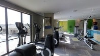 This fitness center offers a thoughtfully designed workout environment spread across a single level. The space is outfitted with a comprehensive selection of cardio equipment, including elliptical trainers, treadmills, and stationary bikes, arranged to maximize floor space and ease of movement between machines. Large windows along one wall flood the interior with natural light, creating an energizing atmosphere while offering views of the surrounding area. The flooring features patterned carpet tiles that add a modern, commercial aesthetic while providing comfort underfoot. Recessed lighting supplements the natural light, ensuring the space remains well-lit throughout. A standout design element is the bold green accent wall at the far end of the room, which adds a vibrant, motivating pop of color and contrasts effectively with the neutral taupe-toned walls. A framed piece of artwork adds a refined touch to the space. The overall ambiance is clean, modern, and purposeful, balancing functionality with contemporary design to create an inviting environment for exercise.