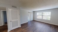 This single-story residence offers a straightforward, functional layout distributed across a series of well-defined spaces. The living area serves as the central gathering point, finished with rich, dark hardwood-style flooring that extends throughout the main areas. Two significant sources of natural light define this room: a large sliding glass door fitted with vertical blinds that provides outdoor access, and an adjacent picture window overlooking greenery outside. A textured popcorn ceiling and neutral gray walls give the space a clean, understated character.

The kitchen is efficiently arranged in an L-shaped configuration, featuring white cabinetry with raised panel detailing, light countertops, a stainless steel refrigerator, a black range, a dishwasher, and a single-basin sink. Light ceramic tile flooring keeps the space practical and easy to maintain.

The bedrooms are carpeted with a warm-toned, plush carpet and share the same neutral gray wall color found throughout the unit. Each bedroom includes at least one window with horizontal blinds, allowing for natural light and outdoor views of surrounding trees. The textured ceilings continue into the bedrooms, maintaining a consistent aesthetic. A separate room with dark hardwood-style flooring, visible in the floor plan model, appears to function as an additional bedroom or flexible space. Overall, the unit presents a clean, move-in-ready environment with cohesive finishes throughout.