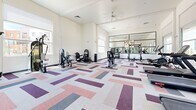 Located at 1115 E Florence Blvd in Casa Grande, AZ, this fitness center offers a well-appointed exercise space designed to support a wide range of workout routines. The single-floor layout is bright and open, with high ceilings and large windows along multiple walls that flood the room with natural light and provide views of the surrounding community.

The exercise room is equipped with a variety of cardio machines, including multiple treadmills and upright stationary bikes, as well as strength-training equipment such as a leg press machine, a weight bench, and additional resistance machines. A full-length mirrored wall on one side enhances the sense of space and allows users to monitor their form during workouts. A ceiling fan provides additional air circulation throughout the room.

The flooring features a bold, geometric patterned carpet in shades of rose, purple, gray, and white, adding a dynamic visual element to the otherwise clean and minimalist interior. White walls and high ceilings contribute to an airy, modern ambiance. A glass door provides direct access to the outdoor area, further connecting the interior to the broader community environment. The space is thoughtfully designed to be both functional and inviting for daily fitness use.