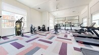 Located at 1161 E O'Neil Dr in Casa Grande, Arizona, this fitness center offers a generously sized, single-floor exercise space designed to support a comprehensive workout experience. The room is thoughtfully laid out to accommodate a diverse selection of equipment, including multiple treadmills, stationary bikes, cable machines, a weight bench, and additional strength-training apparatus arranged to maximize usable floor space.

Architecturally, the space features elevated ceilings that enhance the sense of openness, complemented by large windows along multiple walls that flood the interior with natural light and provide views of the surrounding community. A ceiling fan aids in air circulation, contributing to a comfortable environment during exercise. The walls are finished in crisp white, maintaining a clean and energizing aesthetic throughout.

A striking geometric-patterned carpet in shades of purple, pink, gray, and white adds visual interest underfoot and defines the space's modern character. Full-length mirrors along one wall visually expand the room while serving a practical purpose for form monitoring. A glass-panel door provides direct exterior access, reinforcing the connection to the outdoor surroundings. Overall, the space presents a bright, well-appointed fitness environment suited for a range of training activities.