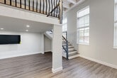 Located at 24 Mt Vernon St in Lynn, MA, this modern loft unit offers a thoughtfully designed open-concept layout with impressive double-height ceilings and an abundance of natural light streaming through large windows. The main living area features smooth hardwood-style flooring and a dramatic staircase with decorative wrought-iron railings leading to an upper loft level, which overlooks the main floor below via a stylish open balcony. Exposed ceiling beams and industrial-style ductwork add authentic character to the space, blending raw architectural detail with clean, contemporary finishes. The kitchen is a standout feature, equipped with stainless steel appliances, white cabinetry, a subway tile backsplash, and a bold navy blue island with dark granite countertops — all set beneath soaring ceilings framed by large street-facing windows. A hallway off the main living area leads to the full bathroom, which includes a tub-shower combo with white tile surround and slate-toned floor tiles. The unit's crisp white walls, recessed and track lighting, and cohesive material palette create a bright, modern ambiance throughout, making this loft a distinctive and functional urban living space.