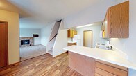 This two-story residence offers a practical and comfortable layout spread across two levels, connected by a carpeted staircase visible from the main living area. The main floor features an open-concept design that integrates the kitchen, dining space, and living room, creating a cohesive and functional flow.

The kitchen is equipped with warm wood-toned cabinetry, a white refrigerator, and a white electric range. A peninsula with a white laminate countertop extends into the space, providing additional prep surface and serving as a natural divider between the kitchen and the adjoining areas. Under-cabinet lighting adds a practical touch above the workspace. A door off the kitchen leads to an additional utility or storage area.

The living room, partially visible from the kitchen, features a fireplace with a stone surround, serving as a focal point and adding warmth to the space. Plush carpet covers the living area, contrasting with the wood-look hard flooring that runs through the kitchen and dining zone. Walls are painted in a neutral light gray, complemented by a textured ceiling throughout. The open sightlines between rooms enhance the sense of space on the main level.