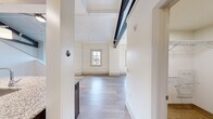 This one-bedroom, one-bathroom loft unit at 338 East 9th Ave in Homestead, PA offers a striking combination of industrial character and modern finishes. The open-concept layout is anchored by soaring ceilings accented with bold, exposed dark steel beams that lend an authentic loft aesthetic throughout. Light hardwood-style flooring runs continuously across the main living areas, enhancing the sense of openness and flow.

The kitchen is well-appointed with rich dark wood cabinetry, gleaming granite countertops, stainless steel appliances including a refrigerator, electric range, and built-in microwave, and a large peninsula island with pendant lighting overhead — ideal for both meal prep and casual dining.

The living area benefits from generous natural light streaming through multiple large windows that frame views of the surrounding Homestead neighborhood and hillside scenery. An interior staircase leads to the loft level, adding vertical dimension to the space.

The bathroom features classic white octagonal tile flooring, a dark wood vanity with clean white countertop, a framed mirror, and updated brushed nickel fixtures. A dedicated in-unit laundry closet houses a stacked washer and dryer for added convenience. Clean white walls throughout keep the space bright and versatile.
