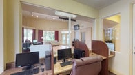 This single-floor facility combines a dedicated business center with an adjoining lounge and common area, finished throughout in warm yellow tones that lend a welcoming atmosphere. The business center section is equipped with multiple desktop computer workstations arranged along granite-topped counters with dark wood privacy dividers, leather rolling chairs, and a shared laser printer. Recessed track lighting illuminates the workspace, while large windows overlook an outdoor pool area and palm-lined grounds, allowing generous natural light into the room. Carpet flooring anchors the workstation area, providing a practical contrast to the hard flooring found in adjacent spaces. A glass partition and open pass-through connect the business center to the broader lounge area, which features a seating arrangement with sofas, hard-surface flooring, and floor-to-ceiling windows dressed in deep red curtains. French doors at the far end of the lounge open to the exterior, enhancing the sense of openness and connectivity to the outdoor environment. A wall-mounted television is positioned for visibility from both zones. The layout flows efficiently between functional and social spaces, making the facility well-suited for both productivity and relaxed gathering.