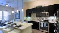 This single-floor residence showcases a thoughtfully designed open-concept layout that blends style and functionality. The kitchen is a standout feature, outfitted with bold dark cabinetry that contrasts beautifully against light quartz countertops and a striking mosaic tile backsplash. Stainless steel appliances, including a range, microwave, and dishwasher, provide a polished, modern aesthetic. A breakfast bar with pendant lighting overhead creates an inviting casual dining area, while under-cabinet lighting adds warmth and practicality to the workspace. The kitchen flows effortlessly into the living area, which is bright and airy, benefiting from an open sightline that enhances the sense of space throughout. Warm wood-look flooring runs throughout the main areas, grounding the interior with a natural, cohesive feel. The neutral wall tones allow the darker cabinetry and accent décor to take center stage. Pendant lights hang at varying heights above the breakfast bar, adding a contemporary design touch that draws the eye upward. The overall ambiance is modern and refined, combining high-end finishes with a welcoming, livable atmosphere that is well-suited for both everyday living and entertaining.