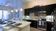 This single-floor residence showcases a thoughtfully designed open-concept layout that blends style and functionality. The kitchen is a standout feature, outfitted with bold dark cabinetry that contrasts beautifully against light quartz countertops and a striking mosaic tile backsplash. Stainless steel appliances, including a range, microwave, and dishwasher, provide a polished, modern aesthetic. A breakfast bar with pendant lighting overhead creates an inviting casual dining area, while under-cabinet lighting adds warmth and practicality to the workspace. The kitchen flows effortlessly into the living area, which is bright and airy, benefiting from an open sightline that enhances the sense of space throughout. Warm wood-look flooring runs throughout the main areas, grounding the interior with a natural, cohesive feel. The neutral wall tones allow the darker cabinetry and accent décor to take center stage. Pendant lights hang at varying heights above the breakfast bar, adding a contemporary design touch that draws the eye upward. The overall ambiance is modern and refined, combining high-end finishes with a welcoming, livable atmosphere that is well-suited for both everyday living and entertaining.