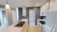 This single-floor space offers a well-designed layout centered around a functional and visually cohesive interior. The kitchen serves as a focal point, featuring white cabinetry with gold-toned trim, a double-basin stainless steel sink set into a white countertop, and white appliances including an electric range and over-the-range microwave. A tiled backsplash near the range adds a subtle decorative touch, while light tile flooring runs throughout, contributing to a bright and clean atmosphere. A pendant light hangs above the counter, providing targeted task lighting, while recessed ceiling lights add to the overall illumination. Bold slate-gray accent walls provide a striking contrast against the white cabinetry and trim, lending the space a modern, intentional design sensibility. An open passageway connects the kitchen to an adjacent carpeted room, which is visible through the opening and illuminated by a chandelier, suggesting a dining or living area. The layout flows naturally between spaces, and the combination of neutral tones, contemporary fixtures, and thoughtful lighting creates a cohesive and inviting environment throughout this single-level home.