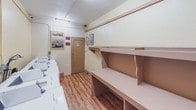 This single-floor laundry facility is designed for practical, shared use and offers a well-organized layout with all the essentials for efficient laundering. A row of white top-load washing machines lines one wall, each equipped with individual controls and coin-operated mechanisms, making the space suitable for multi-unit or communal settings.

Opposite the machines, a series of built-in shelving units provides generous storage and folding space. The tiered shelves, finished in a warm neutral tone, offer multiple levels for sorting, folding, and staging laundry items, complemented by a lower hanging rod for garments. This shelving wall runs nearly the full length of the room, maximizing utility.

The flooring throughout is a warm wood-look laminate that adds a touch of warmth to the otherwise functional space. Cream-colored walls are accented with framed artwork and posted signage, contributing a modest sense of character. Fluorescent lighting fixtures overhead ensure bright, even illumination throughout. A wood-toned door provides access to an adjacent storage closet. The overall ambiance is clean, practical, and neatly maintained.