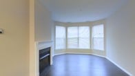 This single-floor space presents a well-appointed living area defined by a combination of classic and contemporary design elements. The room's most distinctive architectural feature is its curved bay window arrangement, where three large windows follow a graceful arc, flooding the interior with abundant natural light and providing views to the outdoors. The windows are fitted with horizontal blinds, allowing for adjustable light control throughout the day.

A traditional white-mantled fireplace anchors one wall of the living area, serving as a focal point that adds both warmth and character to the space. The crisp white surround and classic mantel detailing contrast tastefully against the neutral, warm-toned walls that run throughout the room.

Dark hardwood flooring extends across the entire space, providing a rich, grounding contrast to the light walls and white trim. The smooth, white ceiling with recessed vents contributes to the room's clean, uncluttered aesthetic. Together, these elements create a living space that feels open, airy, and move-in ready, well-suited to a range of furnishing styles.