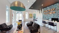 Delray Station's community clubhouse is a thoughtfully designed, single-floor space that blends contemporary style with an inviting, energetic atmosphere. Polished concrete floors extend throughout, reflecting the generous natural light that pours in through floor-to-ceiling glass doors and large multi-pane windows along the perimeter. The soaring ceilings create a grand sense of openness, further elevated by a dramatic oversized dome pendant light finished in brushed gold with a bold teal interior — a standout focal point of the space.

The main lounge area is anchored by tufted charcoal velvet sofas and armchairs arranged around a sleek oval coffee table, offering a comfortable and stylish gathering area. A flat-screen television is mounted nearby, adding a functional amenity to the social setting. The reception desk, backed by an eye-catching mosaic tile accent wall in shades of blue and teal, features the "Delray Station" name rendered in marquee-style lettering, serving as a striking branding element.

Vivid, large-format artwork punctuates the white walls throughout, contributing to the space's vibrant character. Adjacent rooms, visible through open doorways, hint at additional amenity areas with ample daylighting. The overall design is modern and polished, balancing bold aesthetic choices with open, functional layouts.