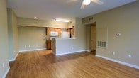 This single-floor unit features an open-concept layout that seamlessly connects the living area and kitchen, creating a functional and cohesive living space. The flooring throughout is a warm-toned hardwood-style material that adds continuity across the main areas.

The living space is generously sized, with neutral beige walls and white baseboards that provide a clean, understated backdrop. A ceiling fan with a light fixture is centrally positioned overhead, offering both ventilation and ambient lighting.

The kitchen is defined by a peninsula-style breakfast bar that separates it from the living area while maintaining the open feel. Cabinetry is finished in a warm medium-wood tone, complementing the overall color palette. White appliances, including a refrigerator, microwave, and range, are built into the layout. A fluorescent light fixture is mounted beneath the upper cabinetry, providing direct task lighting.

A doorway visible from the main area leads to additional rooms, accompanied by a floor-level HVAC vent. Recessed lighting adds supplemental illumination. The overall ambiance is practical and neutral, well-suited for a variety of furnishing styles.