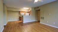 This single-floor unit features an open-concept layout that seamlessly connects the living area and kitchen, creating a functional and cohesive living space. The flooring throughout is a warm-toned hardwood-style material that adds continuity across the main areas.

The living space is generously sized, with neutral beige walls and white baseboards that provide a clean, understated backdrop. A ceiling fan with a light fixture is centrally positioned overhead, offering both ventilation and ambient lighting.

The kitchen is defined by a peninsula-style breakfast bar that separates it from the living area while maintaining the open feel. Cabinetry is finished in a warm medium-wood tone, complementing the overall color palette. White appliances, including a refrigerator, microwave, and range, are built into the layout. A fluorescent light fixture is mounted beneath the upper cabinetry, providing direct task lighting.

A doorway visible from the main area leads to additional rooms, accompanied by a floor-level HVAC vent. Recessed lighting adds supplemental illumination. The overall ambiance is practical and neutral, well-suited for a variety of furnishing styles.