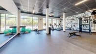 Located at 1370 Stittsville Main St in Ottawa, this single-floor exercise room offers a thoughtfully designed fitness environment that balances functionality with visual energy. The space is anchored by large floor-to-ceiling windows along one wall, flooding the interior with natural light and providing open views of the surrounding greenery and streetscape — an inviting backdrop for cardio workouts.

A standout feature is the bold, hand-drawn-style mural that wraps across two walls, filled with motivational phrases and dynamic illustrated characters, lending the space a vibrant, urban character. The dark suspended tile ceiling contrasts sharply with the clean white walls and structural columns, giving the room a modern, polished aesthetic.

The fitness area is well-appointed with a row of treadmills and stationary bikes positioned to face the windows, along with a cable machine, adjustable weight bench, and a dumbbell rack. Stability balls and additional cardio equipment round out the offerings. The rubber-floored workout zone transitions into a more open, concrete-floored area near the entrance — ideal for stretching or functional training — which also features full-length mirrors and a decorative seating nook. Recessed and linear LED lighting ensures the space is evenly illuminated throughout.