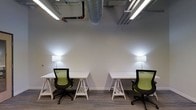This single-floor coworking space offers a clean, modern environment tailored for focused work and collaboration. The interior design embraces an industrial aesthetic, with exposed ductwork, conduit, and piping running along the open ceiling, contrasting beautifully against the smooth white walls and neutral-toned carpet flooring. Overhead, linear LED lighting fixtures provide consistent, bright illumination throughout the space, ensuring a well-lit and energizing atmosphere. Individual workstations are neatly arranged and outfitted with white trestle-style desks and ergonomic mesh-back chairs in a striking green-and-black finish, offering both comfort and style for daily use. Supplemental wall-mounted lamps add a warm, ambient glow at desk level, reducing eye strain and enhancing the overall working environment. The layout is thoughtfully organized to provide each occupant with a defined personal workspace while maintaining an open, airy feel. The combination of industrial ceiling details, minimalist furnishings, and practical lighting makes this space well-suited for professionals, freelancers, or small teams seeking a functional and visually engaging place to work.