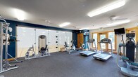 This fitness center offers a comprehensive workout environment designed to accommodate a variety of exercise needs. The space is anchored by a rubberized flooring surface ideal for high-impact activity, and is illuminated by bright fluorescent lighting that ensures excellent visibility throughout. The cardio area is outfitted with multiple treadmills, stationary upright and recumbent bikes, and a rowing machine, providing ample options for aerobic training. Strength training is supported by a dedicated cable and weight machine station. A standout feature is the glass-walled racquetball court adjacent to the main floor, offering an additional athletic amenity visible from the workout area. The walls are finished in a bold navy blue accent color, lending the space a modern, energetic aesthetic. A wall-mounted television keeps members entertained during workouts. Natural light filters in through wood-framed glass doors and windows along one wall, creating a welcoming atmosphere. The facility also includes a ceiling fan for added ventilation and comfort. Exit signage and safety equipment are clearly visible, reflecting a commitment to accessibility and safety standards throughout the space.