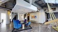 This single-floor space masterfully blends industrial aesthetics with contemporary design, resulting in a bold and energetic environment. The exposed concrete ceiling reveals an intricate network of ductwork and mechanical systems, lending an authentic urban character to the interior. Wide-plank hardwood flooring runs throughout, providing a warm contrast to the raw overhead elements.

The lounge area serves as a striking focal point, anchored by a bold black-and-white geometric area rug. A pair of plush royal blue velvet armchairs and a classic accent chair surround a sculptural side table, creating an inviting seating vignette full of personality. Pendant lighting in varied metallic finishes hangs above, adding a curated, artistic touch.

The walls feature a mix of painted geometric patterns in black and white alongside vibrant, colorful artwork that injects energy and creativity into the space. Concrete accent walls provide textural depth and reinforce the industrial character. A staircase with metal railings is visible in the background, hinting at additional levels or mezzanine access. The overall ambiance is modern, expressive, and thoughtfully designed — ideal for a creative office, coworking hub, or boutique hospitality venue.