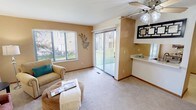 This single-floor residence offers a well-organized layout designed for comfortable everyday living. The living room serves as the central gathering space, finished with plush wall-to-wall carpeting, warm neutral-toned walls, and a ceiling fan overhead. Natural light enters through a large window and a sliding glass door fitted with vertical blinds, which provides direct access to an outdoor patio area with views of a green lawn. A pass-through opening connects the living area to the adjacent kitchen, enhancing the open feel between spaces.

The kitchen is efficiently laid out with white cabinetry featuring wood-tone trim, light countertops, and an electric range. A closet pantry adds practical storage within easy reach.

Two bedrooms are positioned toward the rear of the unit, each carpeted and filled with natural light from windows dressed with blinds and sheer curtains. Both rooms include open closets with shelving for organized storage. The primary bedroom is generously sized and is conveniently connected to the full bathroom, which features a single-sink vanity with wood-framed cabinetry, a lighted mirror, and a shower-tub combination. Throughout the residence, a consistent palette of soft neutrals and warm wood accents creates a calm, cohesive ambiance.