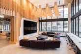 This dynamic single-floor space is a thoughtfully designed collaborative environment that blends open-plan flexibility with distinct functional zones. The main lounge area commands immediate attention with its soaring double-height ceiling, an expansive wall of floor-to-ceiling windows that flood the interior with natural light, and a striking cluster of sculptural pendant lights overhead. Plush sectional sofas and lounge seating create inviting social areas, while a long workbench along the window wall provides dedicated desk space with outdoor views of tree-lined streets.

A kitchen and refreshment station features bold geometric tile backsplash, and is surrounded by vivid, large-scale graphic murals that energize the space with color and artistic character. Exposed concrete ceilings with visible ductwork and bold blue structural columns lend an industrial edge to this area, complementing the warm wood finishes found throughout.

Additional lounge nooks feature fabric sofas, accent chairs, wall-mounted displays, and slatted wood partitions that define spaces without fully enclosing them. A large conference table with rolling chairs offers a formal meeting option, positioned near a branded "Theory" feature wall with neon signage. A monumental multi-panel digital display serves as a focal art installation near the entrance, anchoring the space with a strong visual statement.