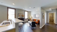 This single-floor space showcases a thoughtfully designed open-concept layout that connects the kitchen and living areas into one cohesive environment. The kitchen features a sleek white breakfast bar with bar stool seating, offering a casual dining option while maintaining a visual connection to the rest of the space. Recessed lighting throughout the ceiling provides consistent, modern illumination, complementing the natural light that filters in through multiple windows dressed with clean white blinds and warm wood trim.

The living area is anchored by a vibrant orange sofa accented with gray throw pillows, paired with a contemporary charcoal armchair and a white rectangular coffee table atop a patterned area rug. Dark hardwood flooring runs throughout, adding warmth and contrast against the neutral gray walls. A media console along one wall provides storage and display space, while framed wall art adds curated character to the room. The overall ambiance is modern and inviting, balancing clean lines with warm color accents for a comfortable yet stylish aesthetic.