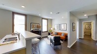 This single-floor space showcases a thoughtfully designed open-concept layout that connects the kitchen and living areas into one cohesive environment. The kitchen features a sleek white breakfast bar with bar stool seating, offering a casual dining option while maintaining a visual connection to the rest of the space. Recessed lighting throughout the ceiling provides consistent, modern illumination, complementing the natural light that filters in through multiple windows dressed with clean white blinds and warm wood trim.

The living area is anchored by a vibrant orange sofa accented with gray throw pillows, paired with a contemporary charcoal armchair and a white rectangular coffee table atop a patterned area rug. Dark hardwood flooring runs throughout, adding warmth and contrast against the neutral gray walls. A media console along one wall provides storage and display space, while framed wall art adds curated character to the room. The overall ambiance is modern and inviting, balancing clean lines with warm color accents for a comfortable yet stylish aesthetic.